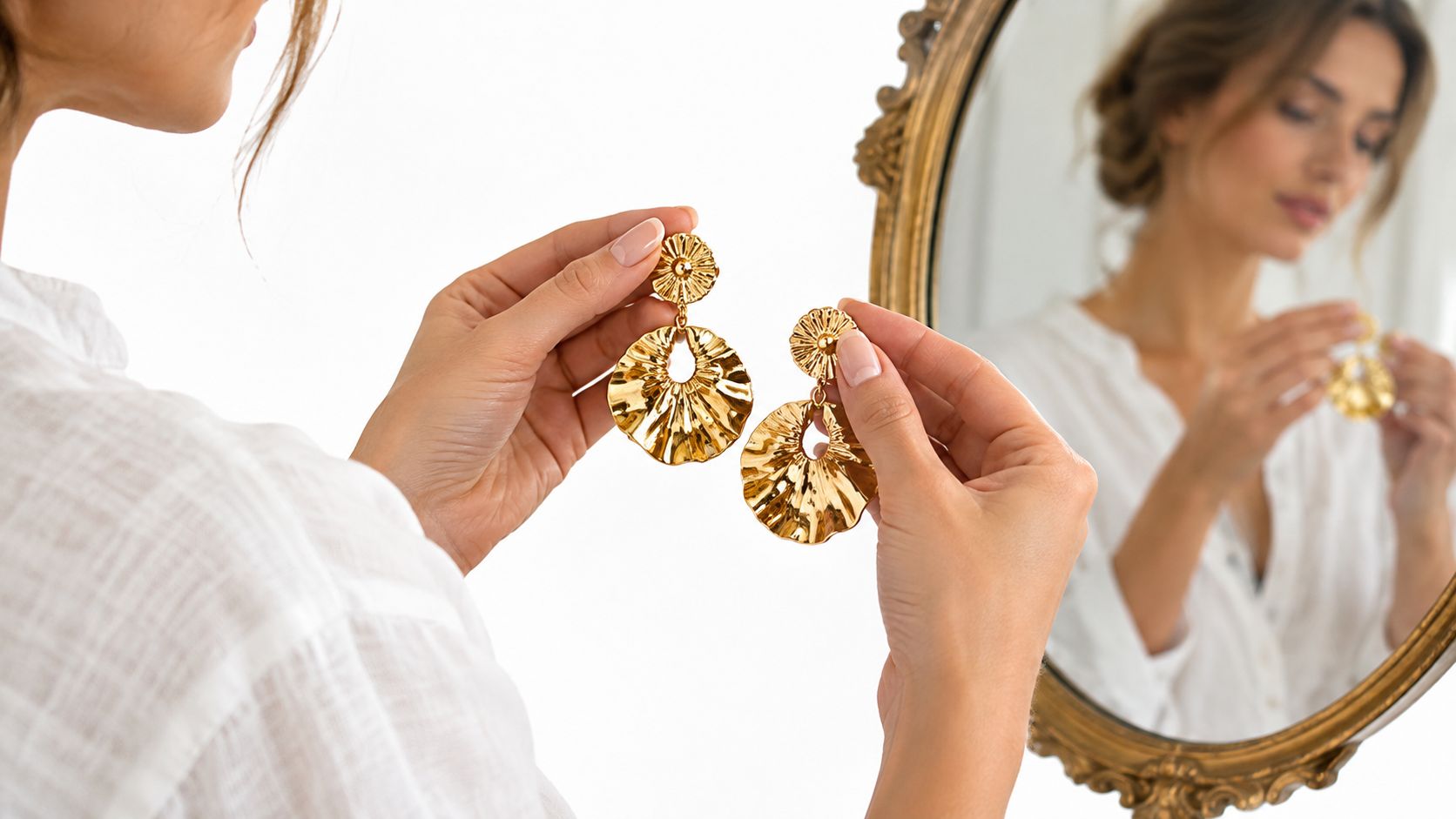 A person holds up elegant gold statement earrings towards a mirror reflecting her face and jewelry.