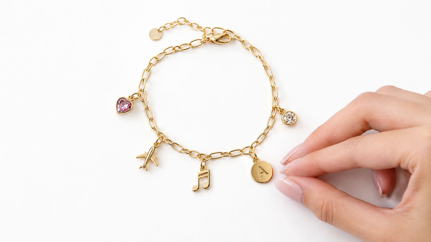 A hand adjusts a delicate gold chain bracelet featuring diverse charms, including a heart, airplane, musical note, and letter A.