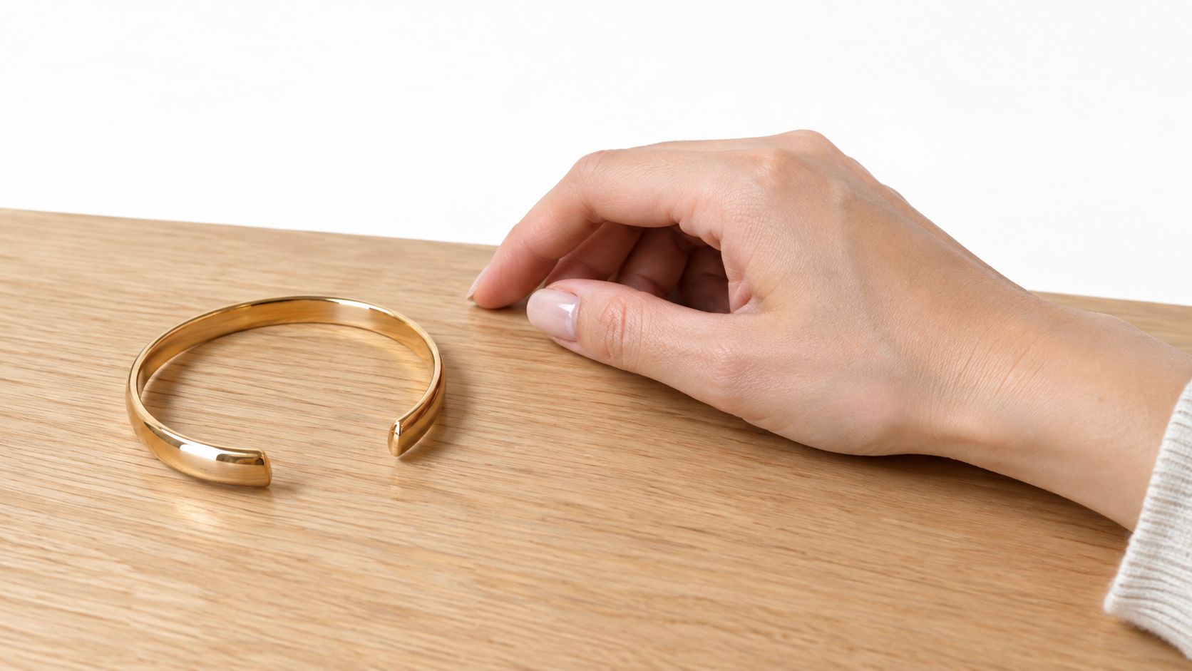 A close-up of a elegant polished gold cuff bracelet sitting on a light oak wooden table beside a hand.