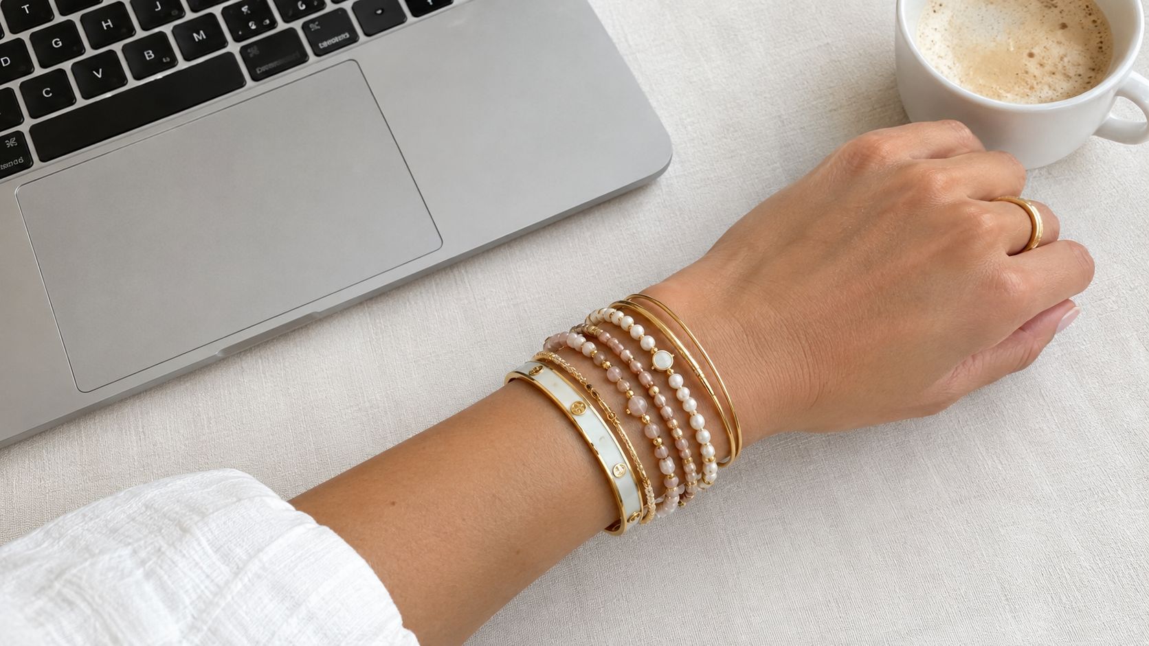 A person's arm decorated with multiple gold and beaded bracelets resting near a laptop and coffee.