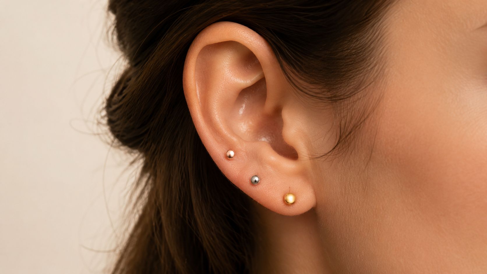 Close-up of a person's ear wearing a minimalist ear stack with three small gold and silver ball earrings.