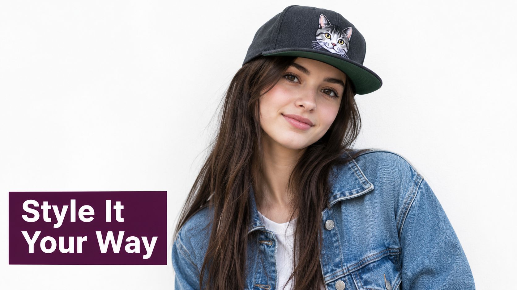 A young woman wearing a denim jacket and a black snapback hat with an embroidered cat face.