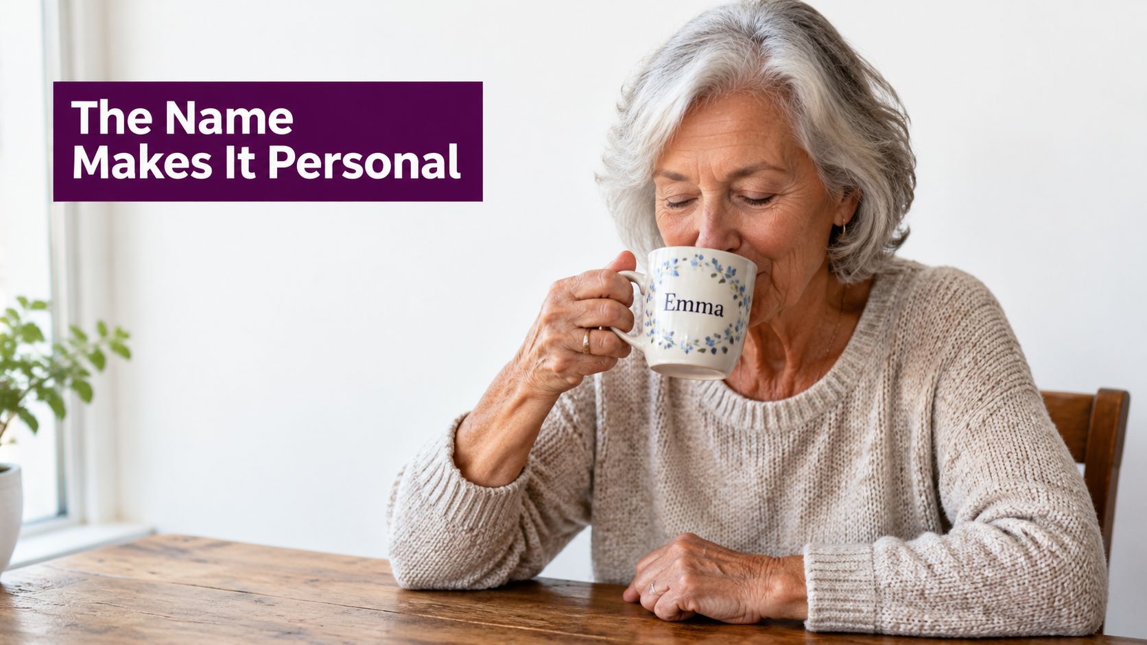 An elderly woman with white hair sitting at a wooden table holding a personalized coffee mug.