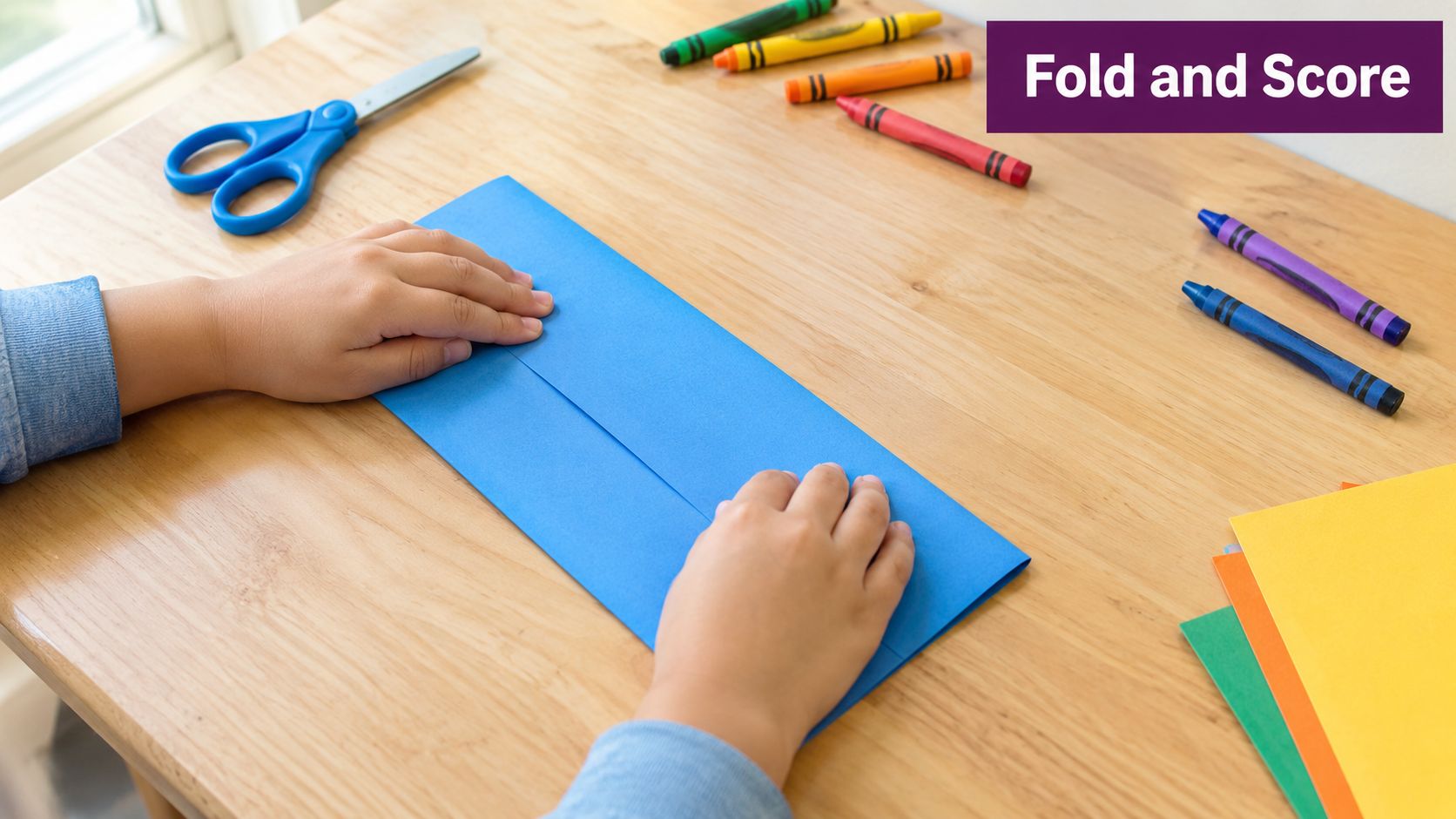 A child's hands carefully folding a blue piece of construction paper on a wooden desk.