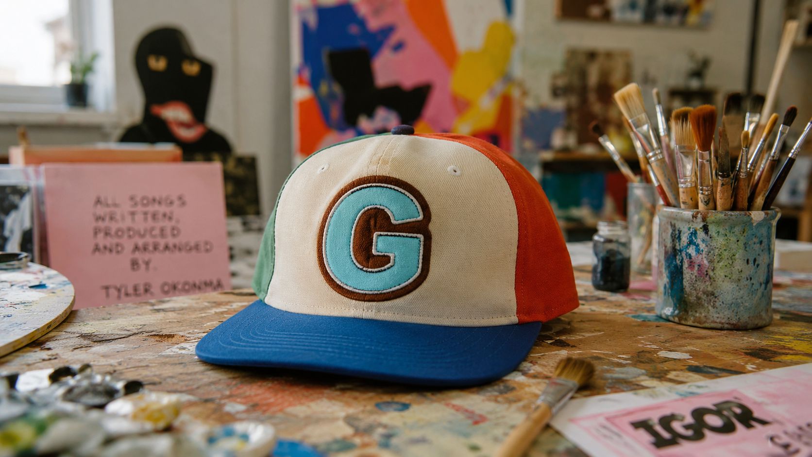 A colorful Golf Wang baseball cap with a prominent G logo sitting on a messy artist workspace.