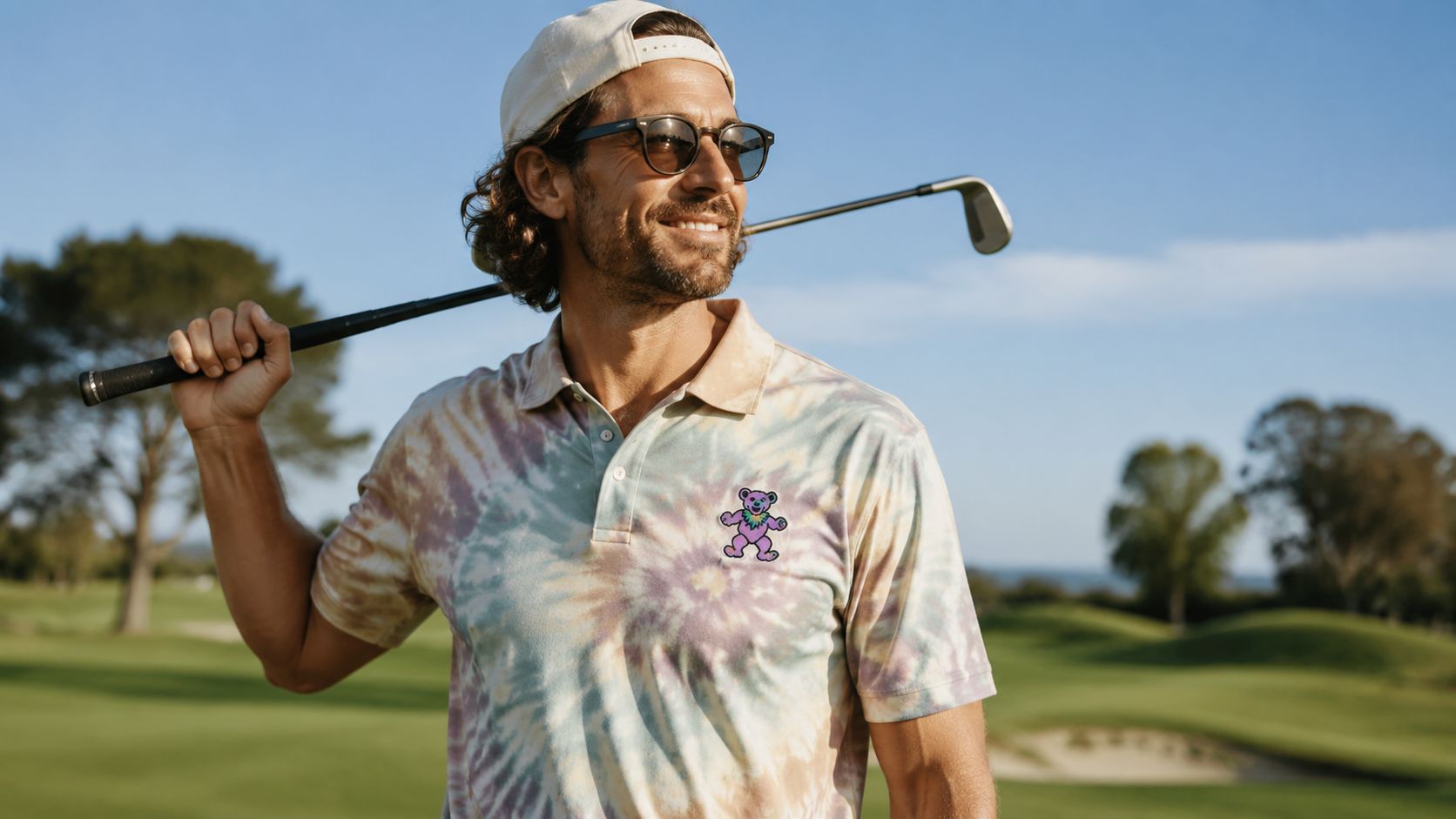 A smiling man wearing a Grateful Dead tie-dye golf shirt and a cap while holding a club.
