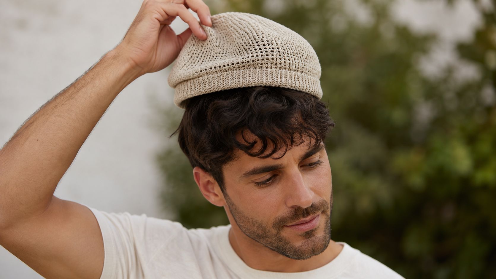 A stylish man with dark curly hair wearing a lightweight beige crochet summer beanie hat outdoors.