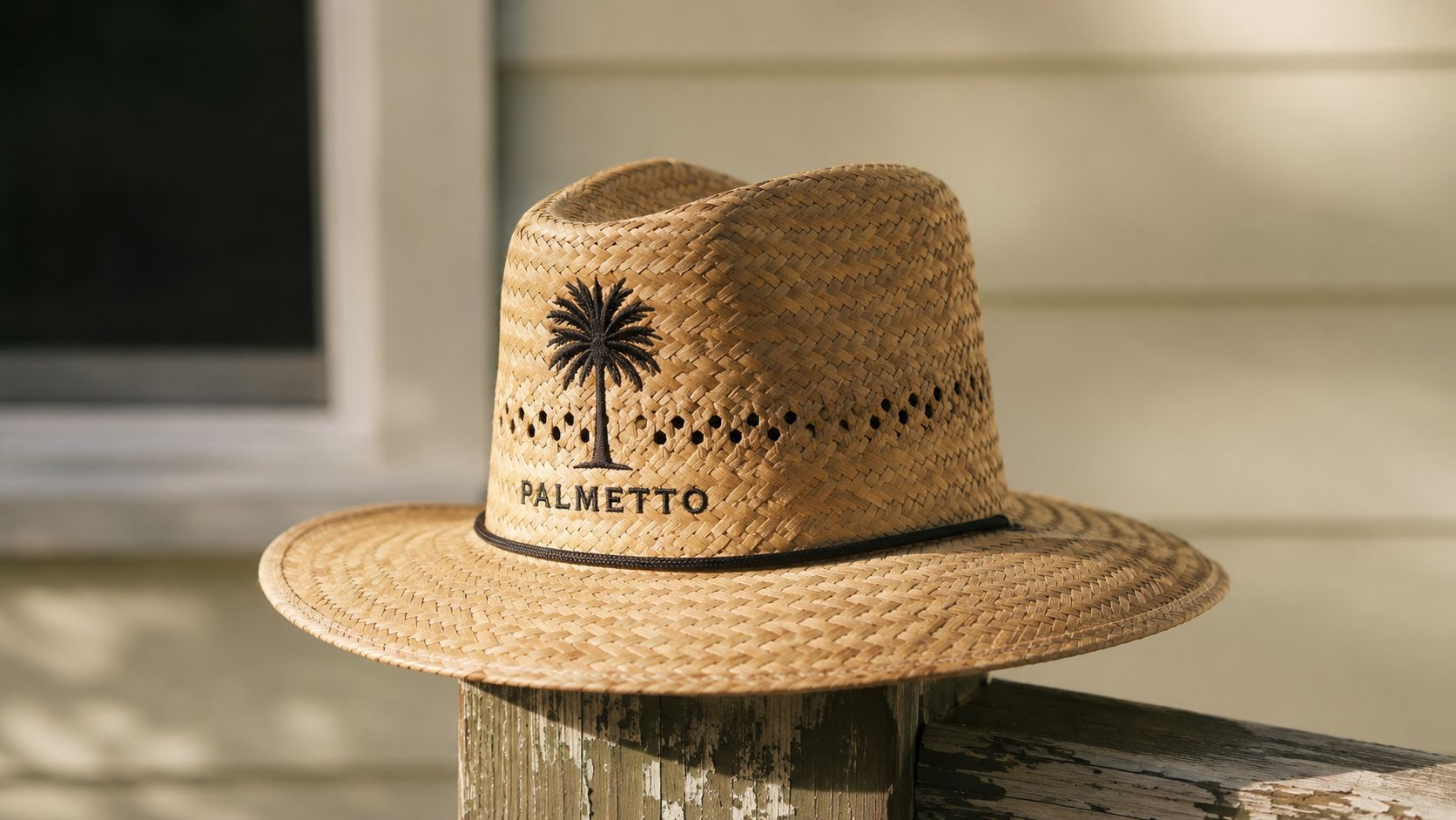 A straw hat with a black palmetto tree emblem and text resting on a wooden porch post.
