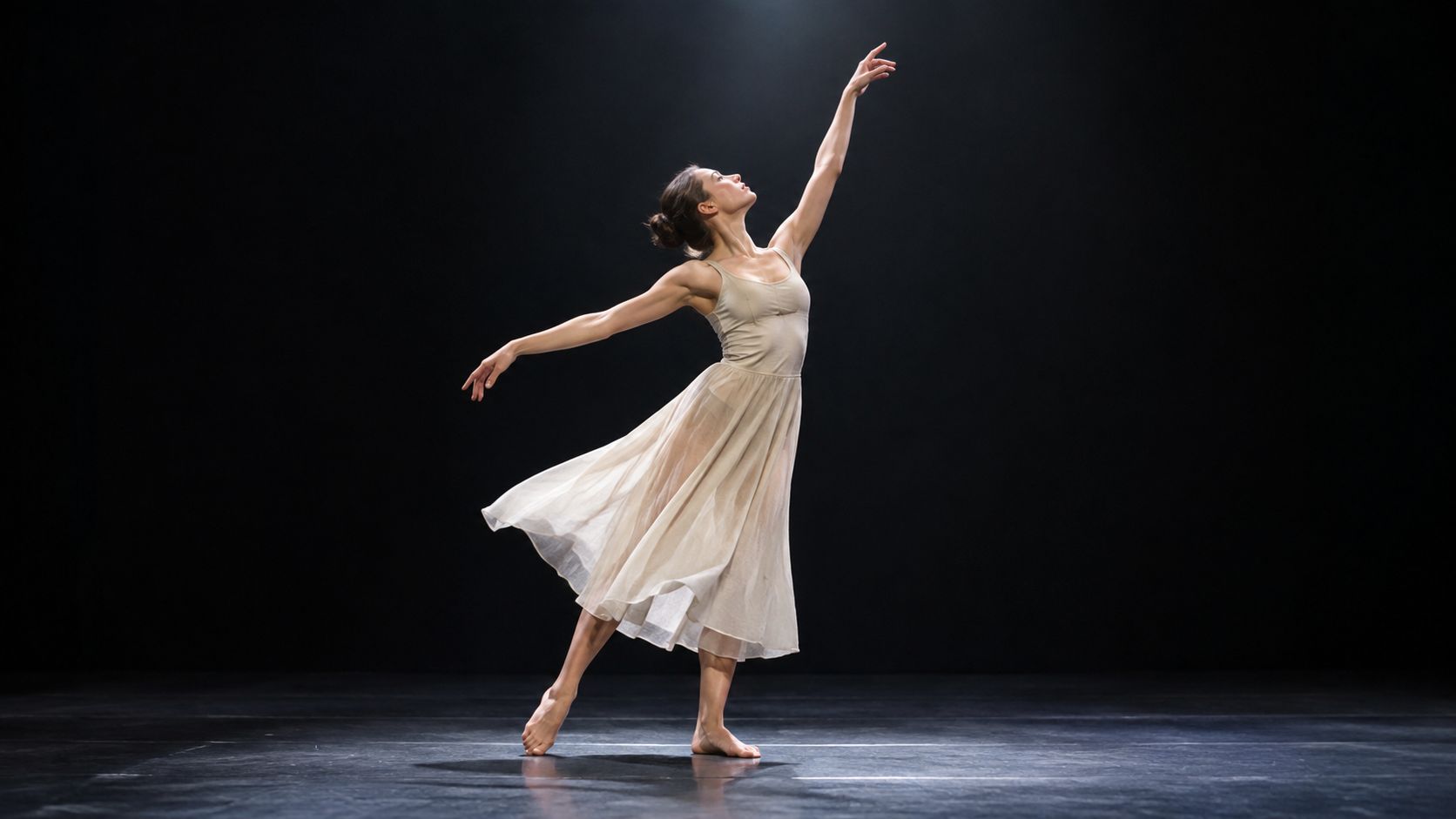 Una bailarina con vestido beige claro realiza una elegante pose de danza contemporánea sobre un escenario oscuro.