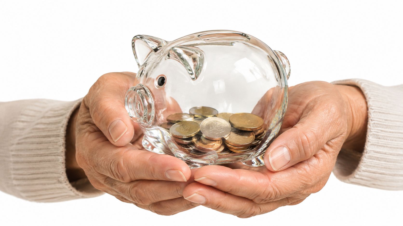 Manos de una persona mayor sosteniendo una hucha de cristal transparente llena con monedas de ahorro.
