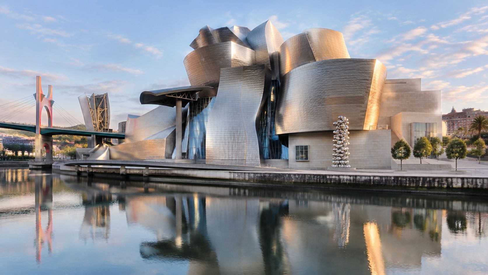 The iconic Guggenheim Museum Bilbao with its unique metallic architecture reflecting on the tranquil river water surface.