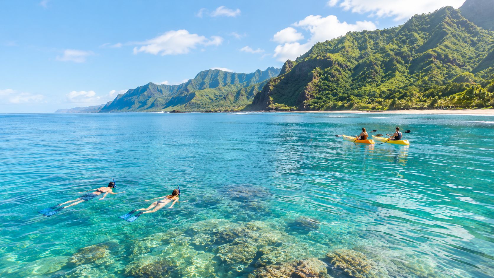 Two people snorkeling in clear tropical water near two kayakers with mountains in the background in Kauai.