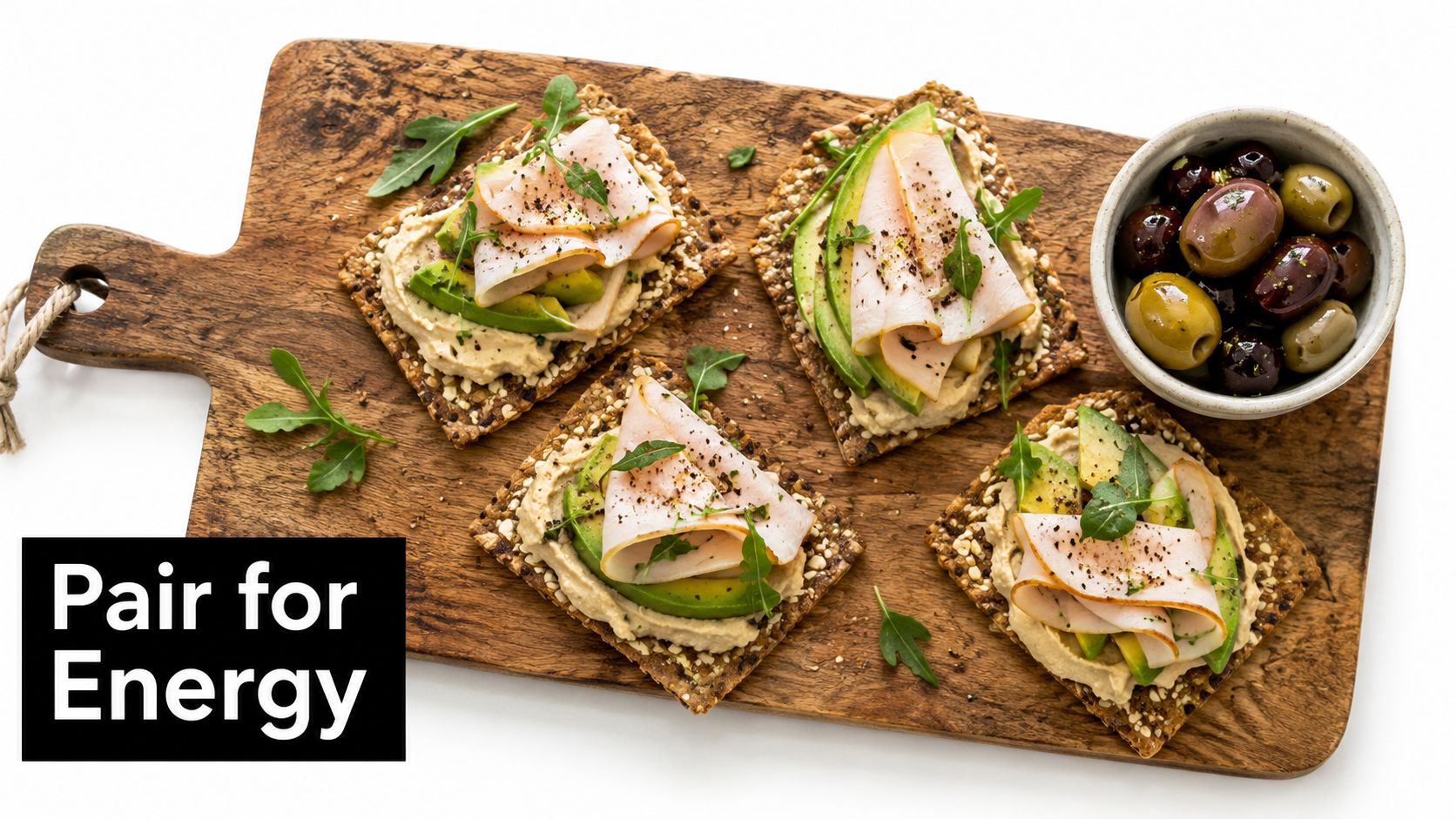 Four crispbread crackers topped with hummus, avocado, and sliced turkey on a wooden cutting board with olives.