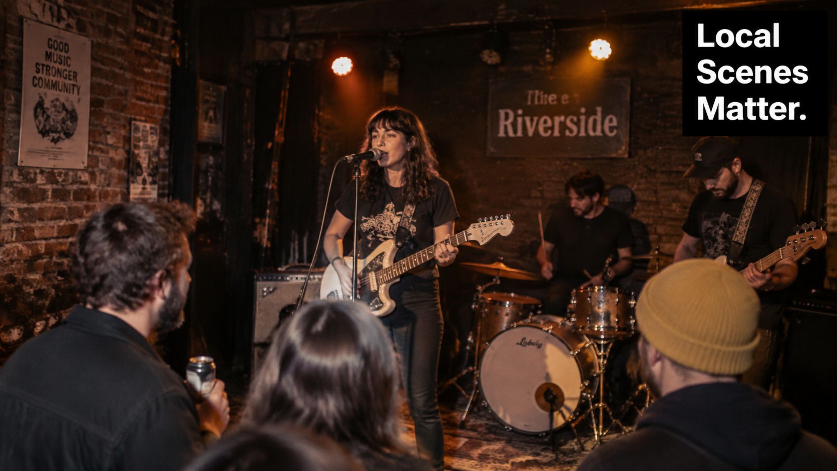 A female musician sings and plays electric guitar with a band at a small, dimly lit venue.