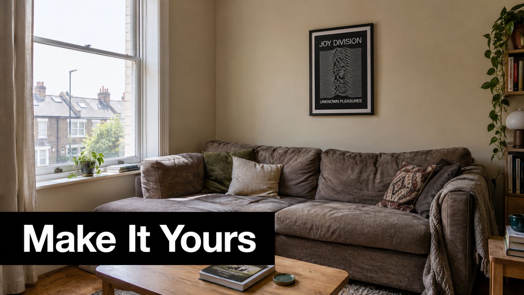 A cozy, sunlit living room featuring a comfortable brown sectional sofa and a Joy Division poster.