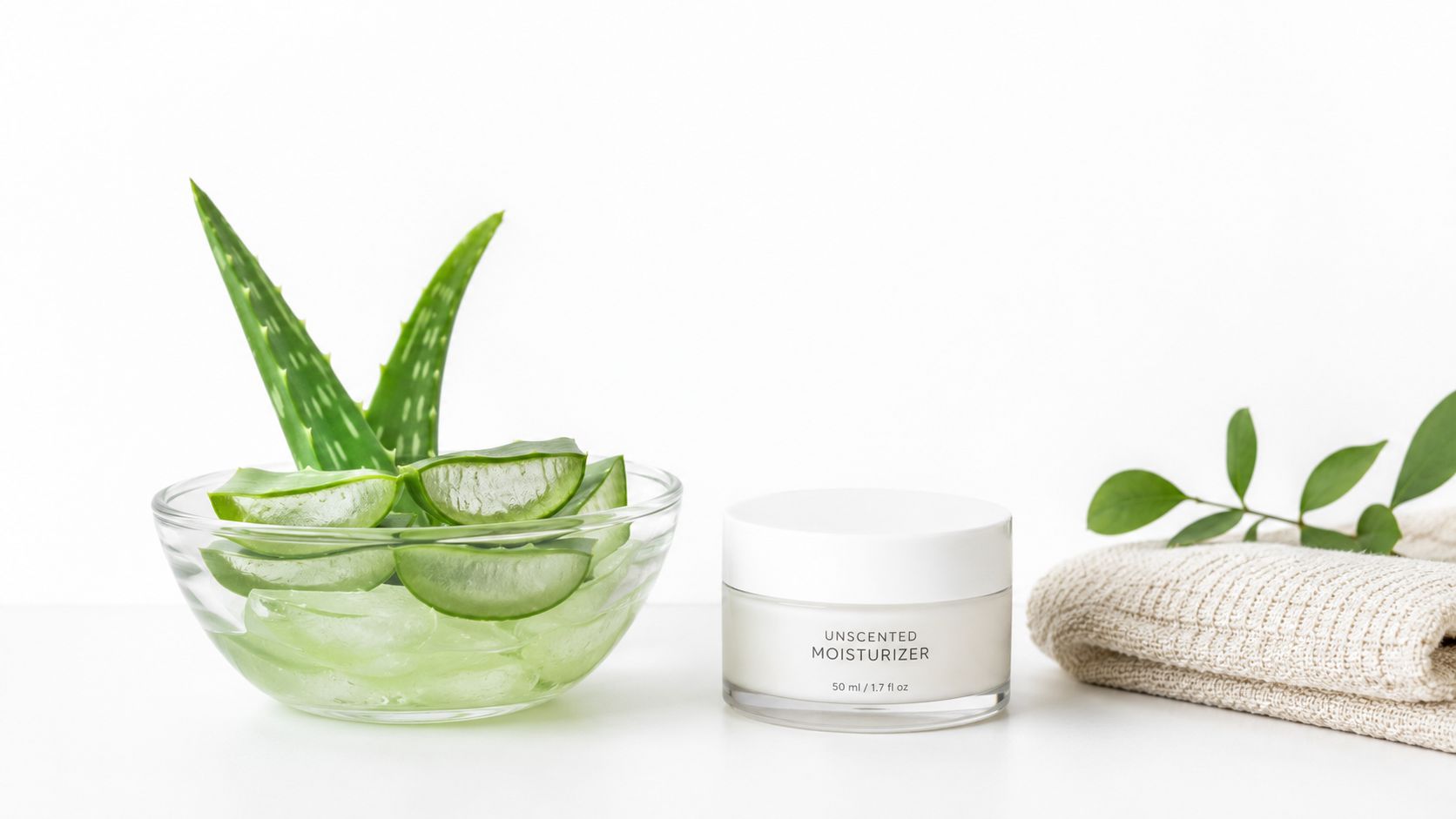 A glass bowl with fresh aloe vera slices next to an unscented moisturizer jar and a towel.