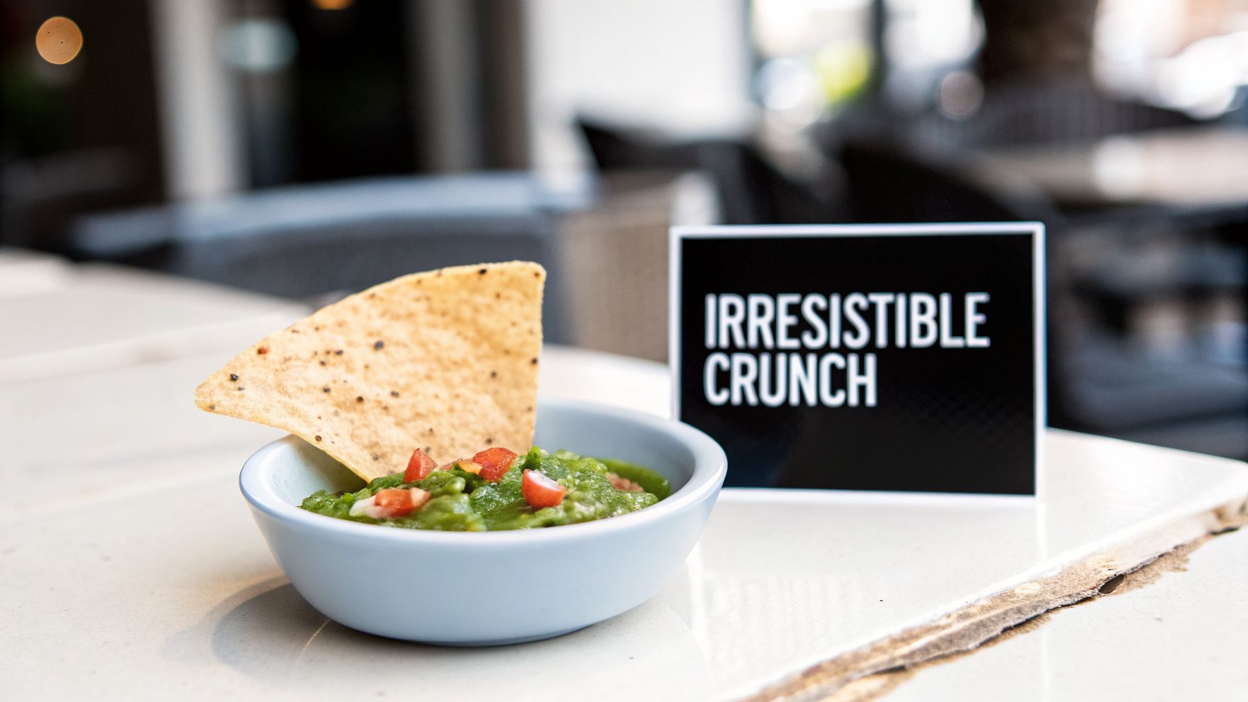 A light blue bowl of fresh green guacamole with red tomatoes, a tortilla chip, and a 'Irresistible Crunch' sign.
