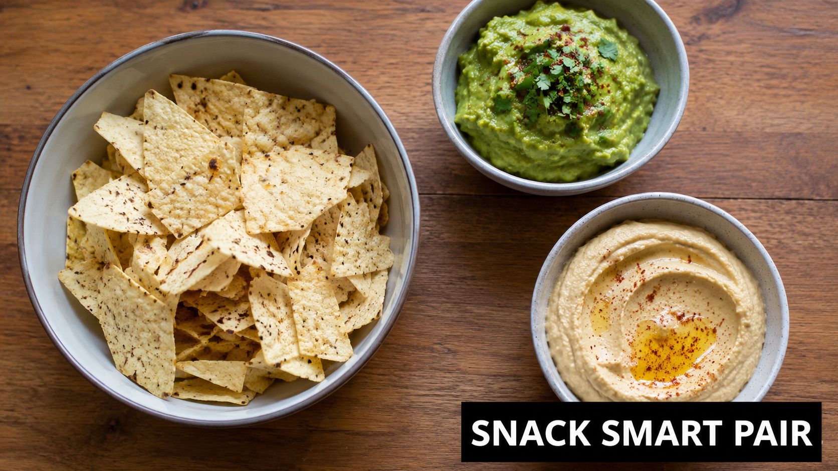 A high-angle shot of a bowl of tortilla chips next to bowls of guacamole and creamy hummus.