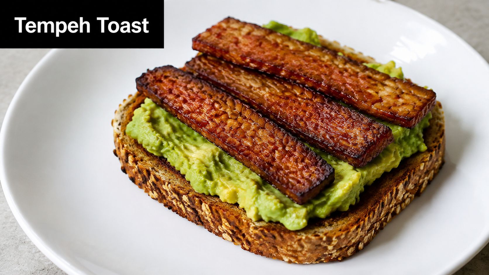 A piece of toasted multigrain bread topped with smashed avocado and strips of pan-seared smoky tempeh strips.