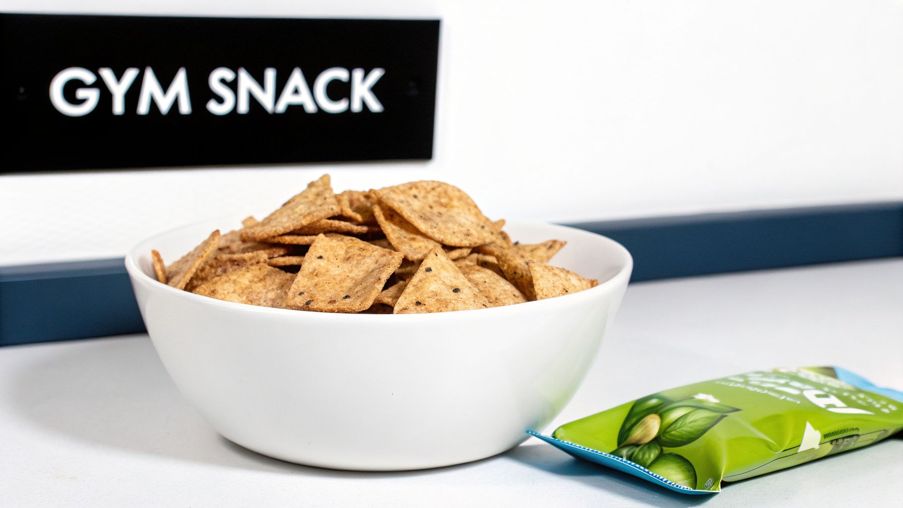 A white bowl overflowing with healthy multigrain tortilla chips, a green snack bag, and a 'GYM SNACK' sign.