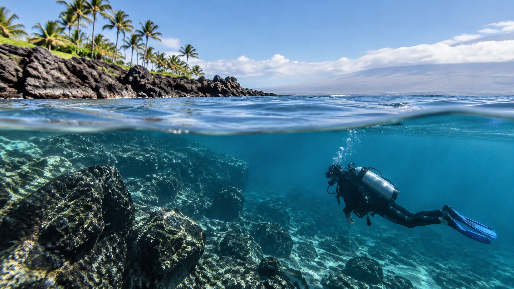 A scuba diver explores the clear blue underwater terrain near a volcanic tropical coastline in Hawaii.