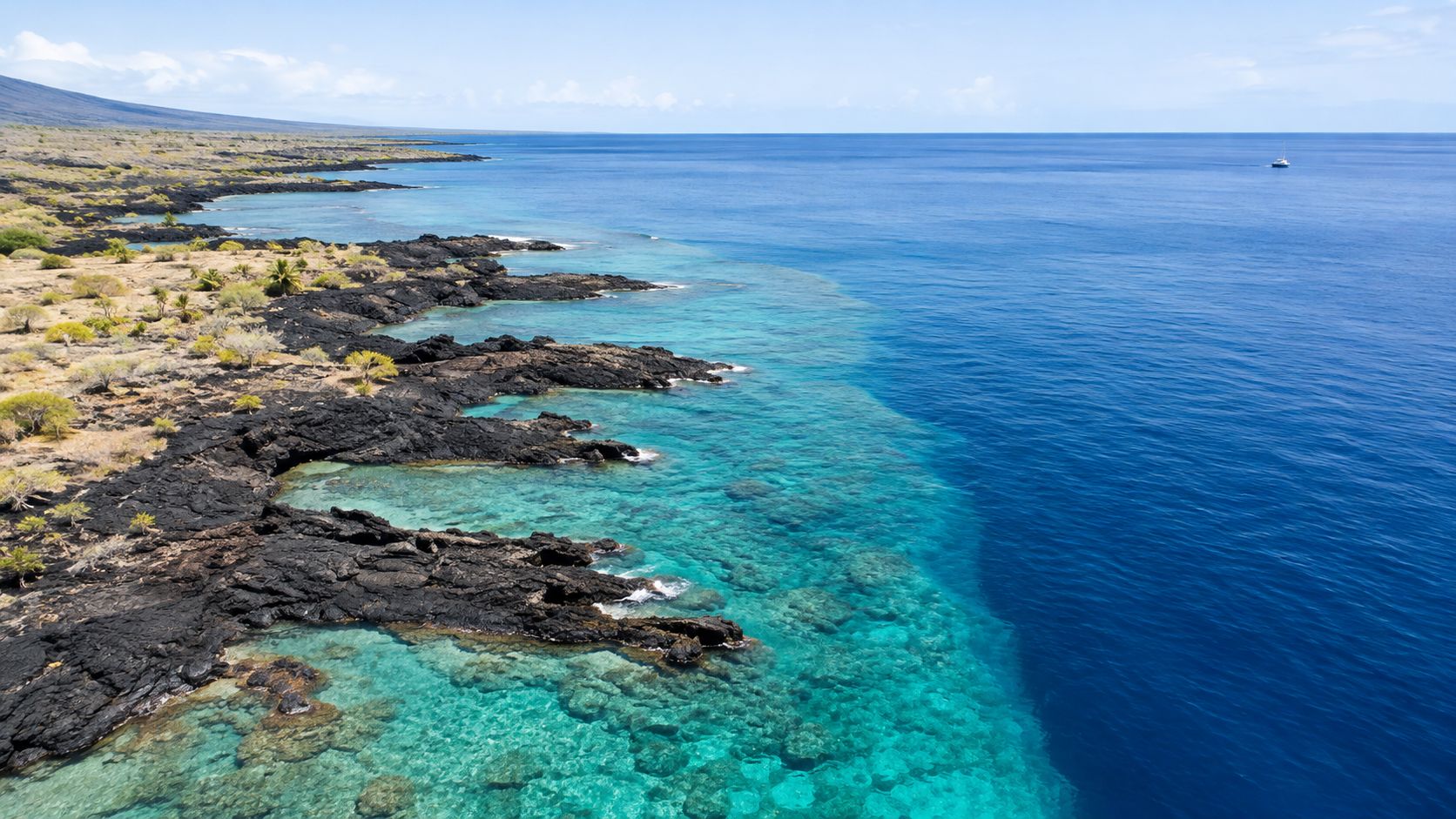 An aerial view of the rugged volcanic coastline with turquoise shallow water meeting the deep blue ocean.