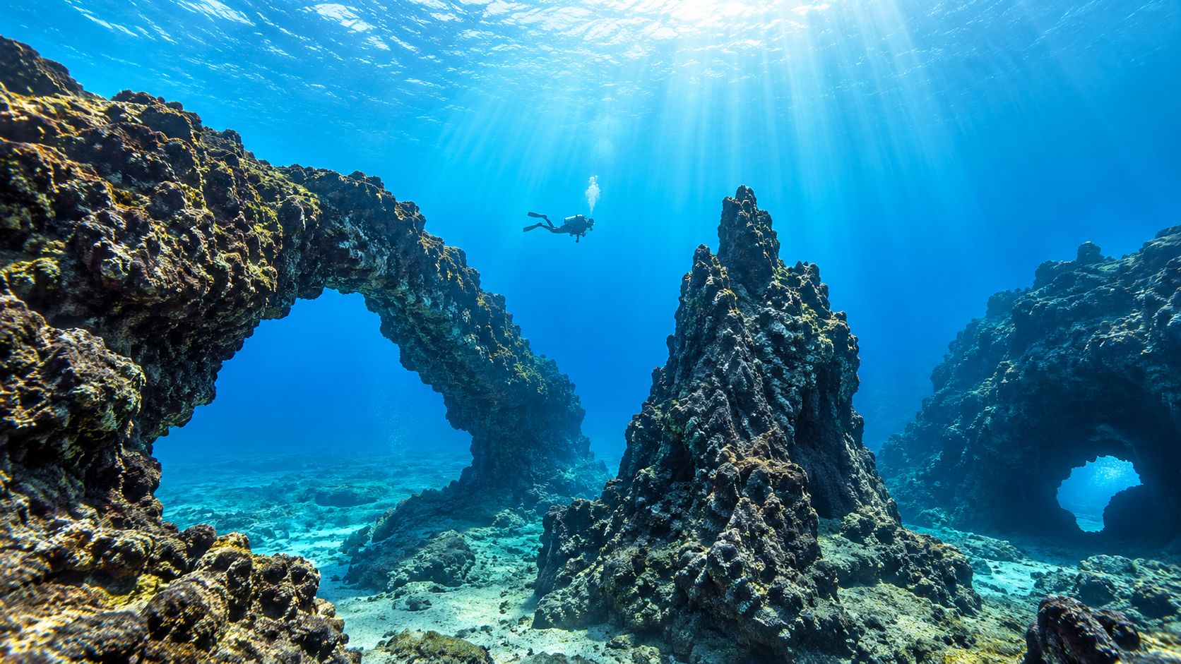 A scuba diver explores a stunning underwater volcanic rock arch formation in clear blue ocean water.