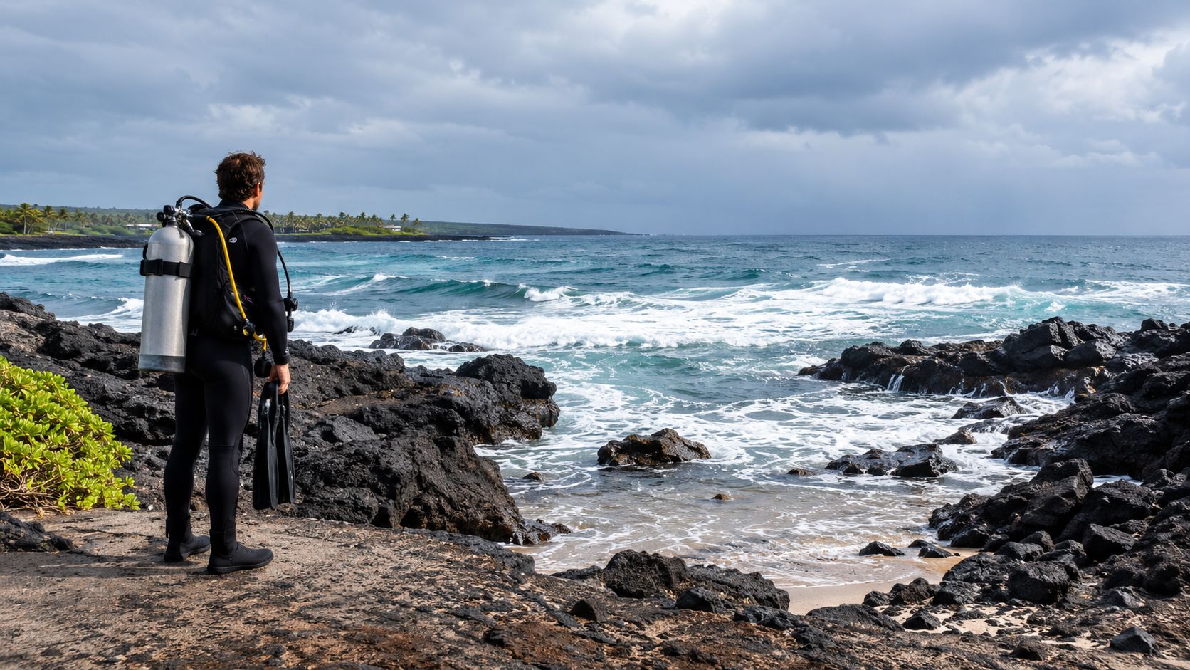 A scuba diver wearing full gear stands on volcanic rocks looking out at the Pacific ocean coast.