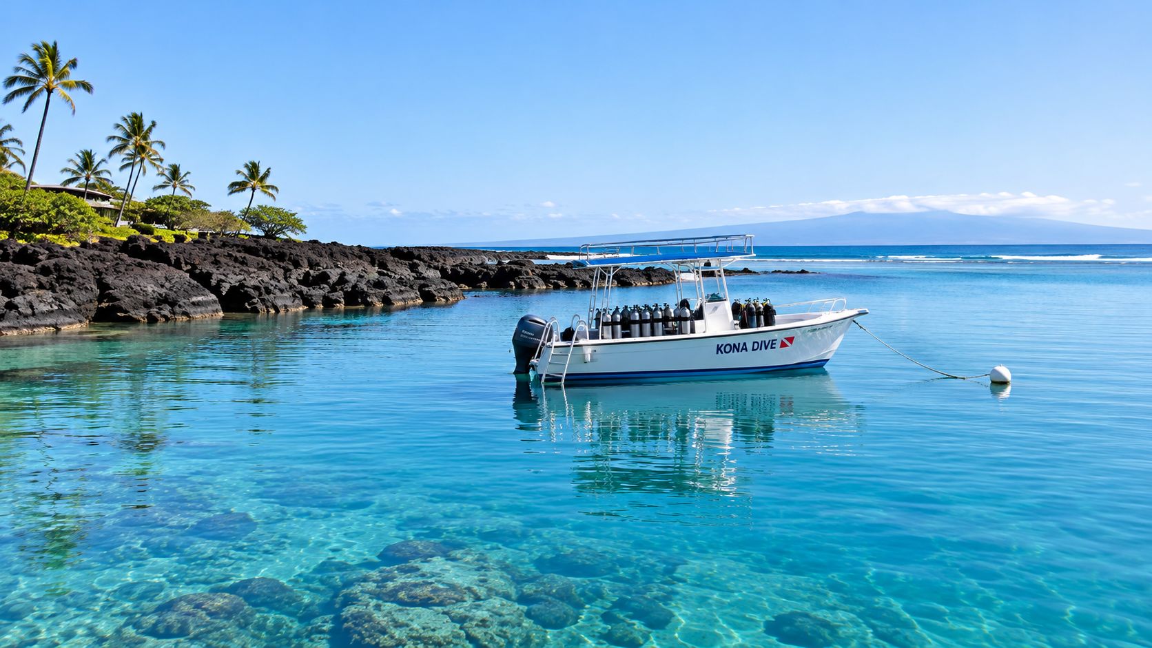 A white dive boat floating in calm, crystal clear turquoise waters near a black volcanic rocky shore.