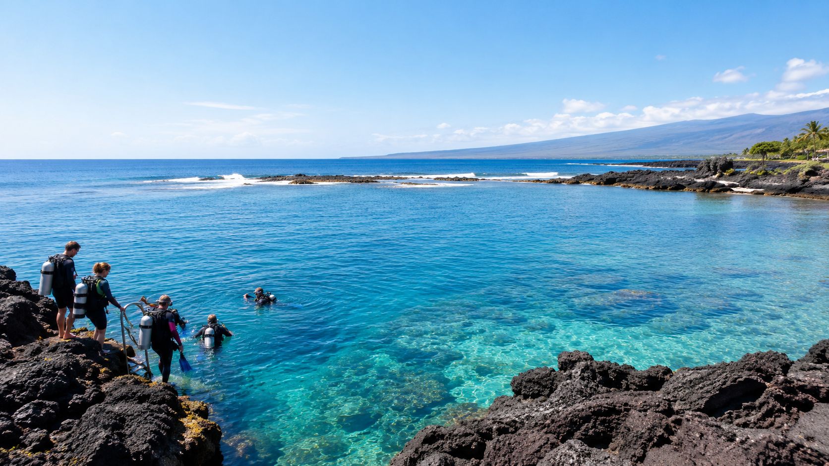 A group of scuba divers prepares to enter the clear blue ocean from a rocky lava shore.