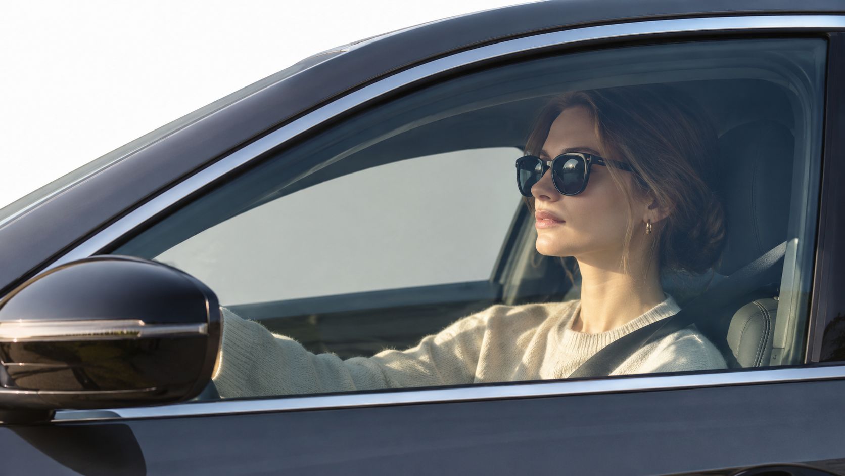 A young woman wearing sunglasses driving a black car while looking forward through the side window.