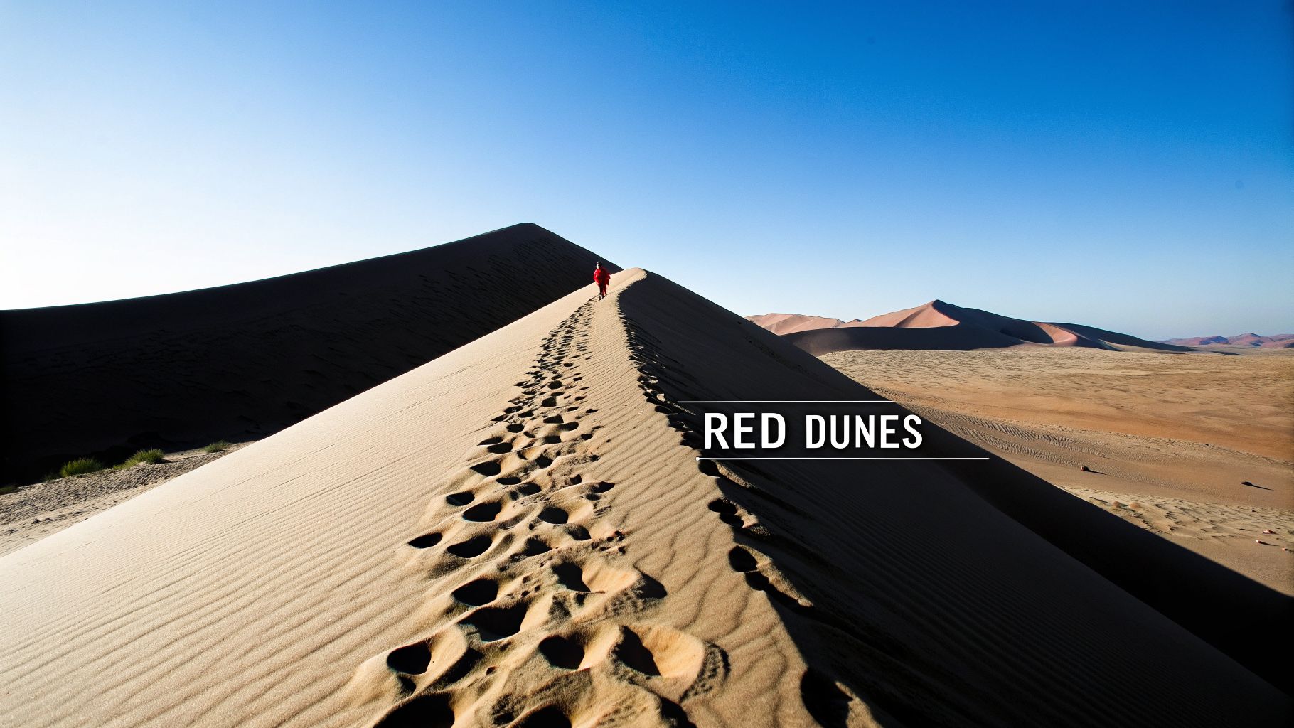 A person in red walks along a sandy red dune, leaving footprints in a vast desert landscape.