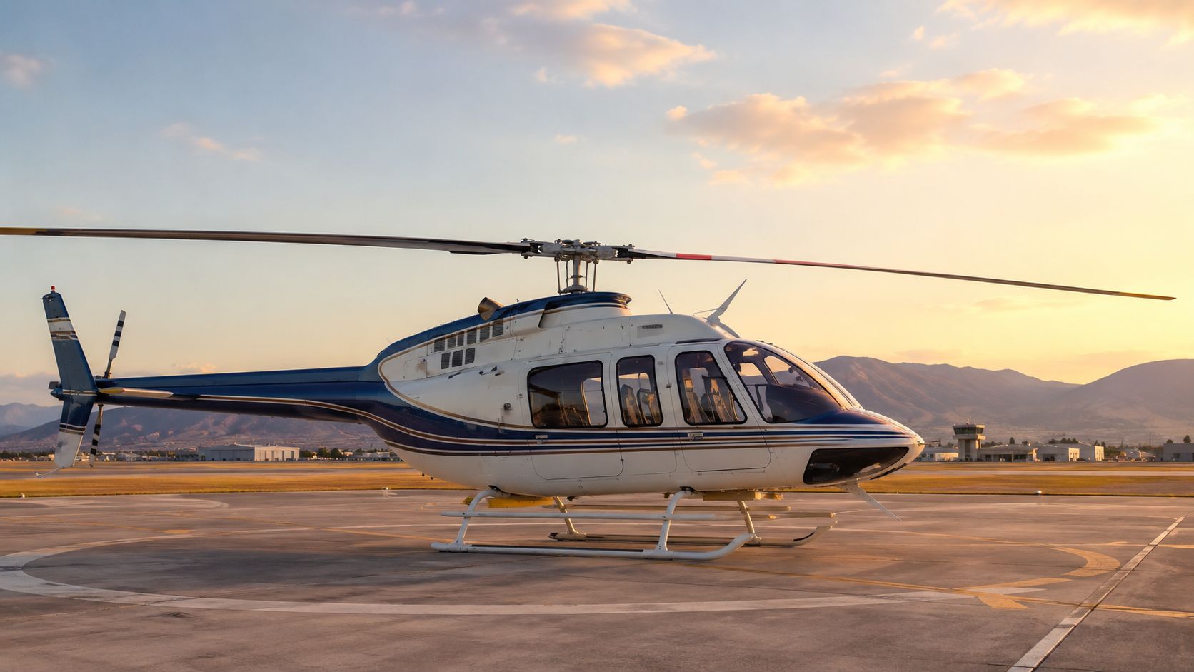 A white and blue Bell 206L4 LongRanger IV helicopter parked on an airport helipad during golden hour.