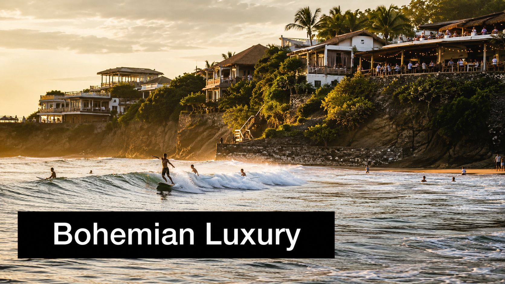People surfing on waves at sunset in front of luxury beachside villas on a rocky cliff.