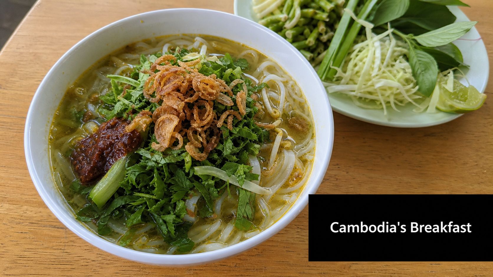 A steaming bowl of Khmer noodle soup with fresh herbs and a side plate of raw vegetables.