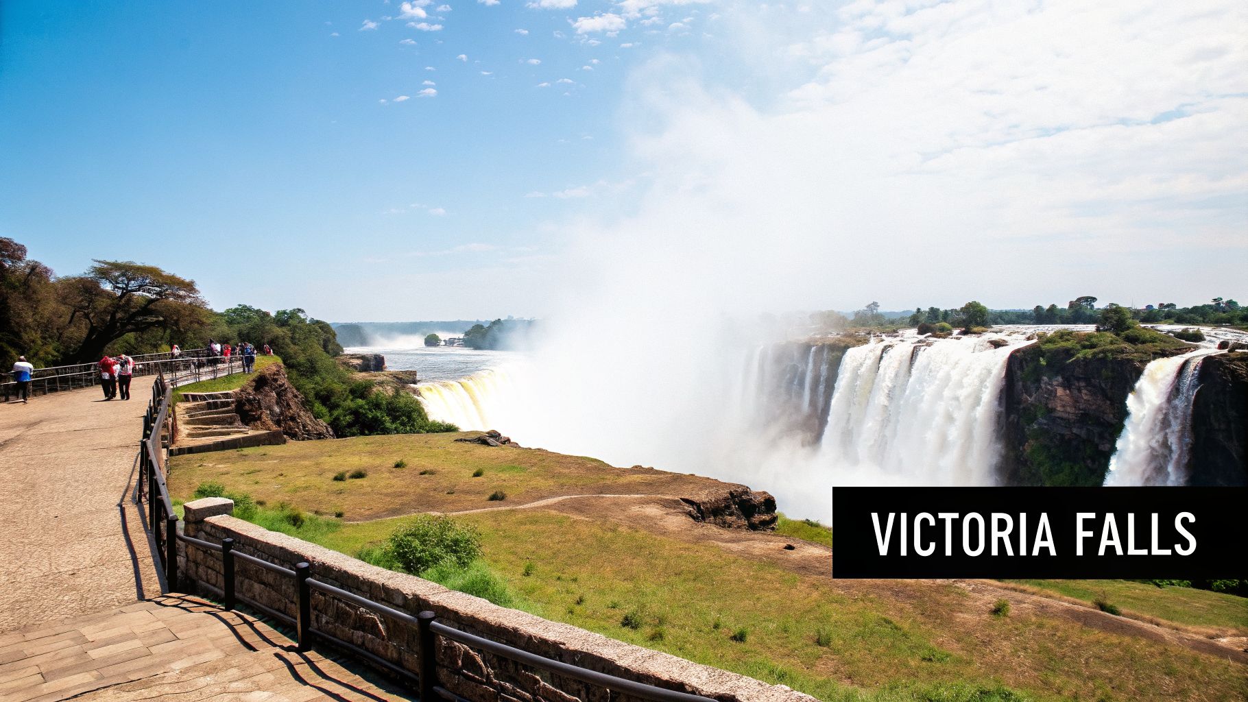 A panoramic view of Victoria Falls showing powerful waterfalls, mist, and tourists on a pathway.