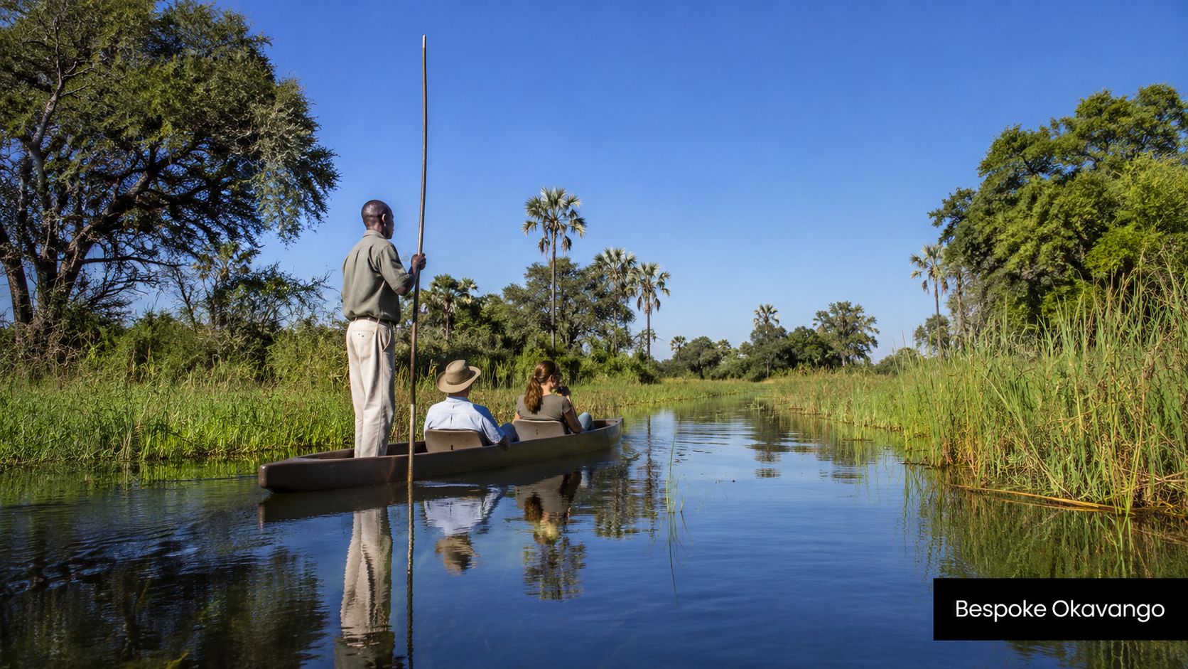 A guide poles a traditional mokoro canoe carrying two tourists through the calm waters of the Okavango Delta.