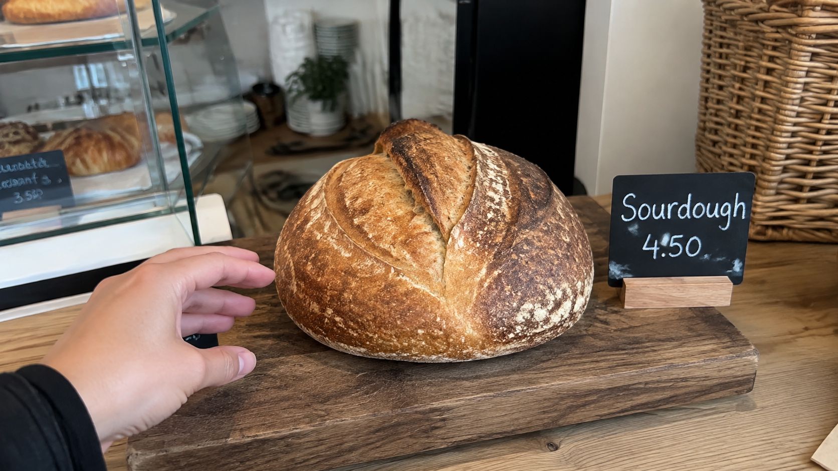 A hand reaching towards a freshly baked crusty round sourdough bread loaf displayed on a wooden counter.