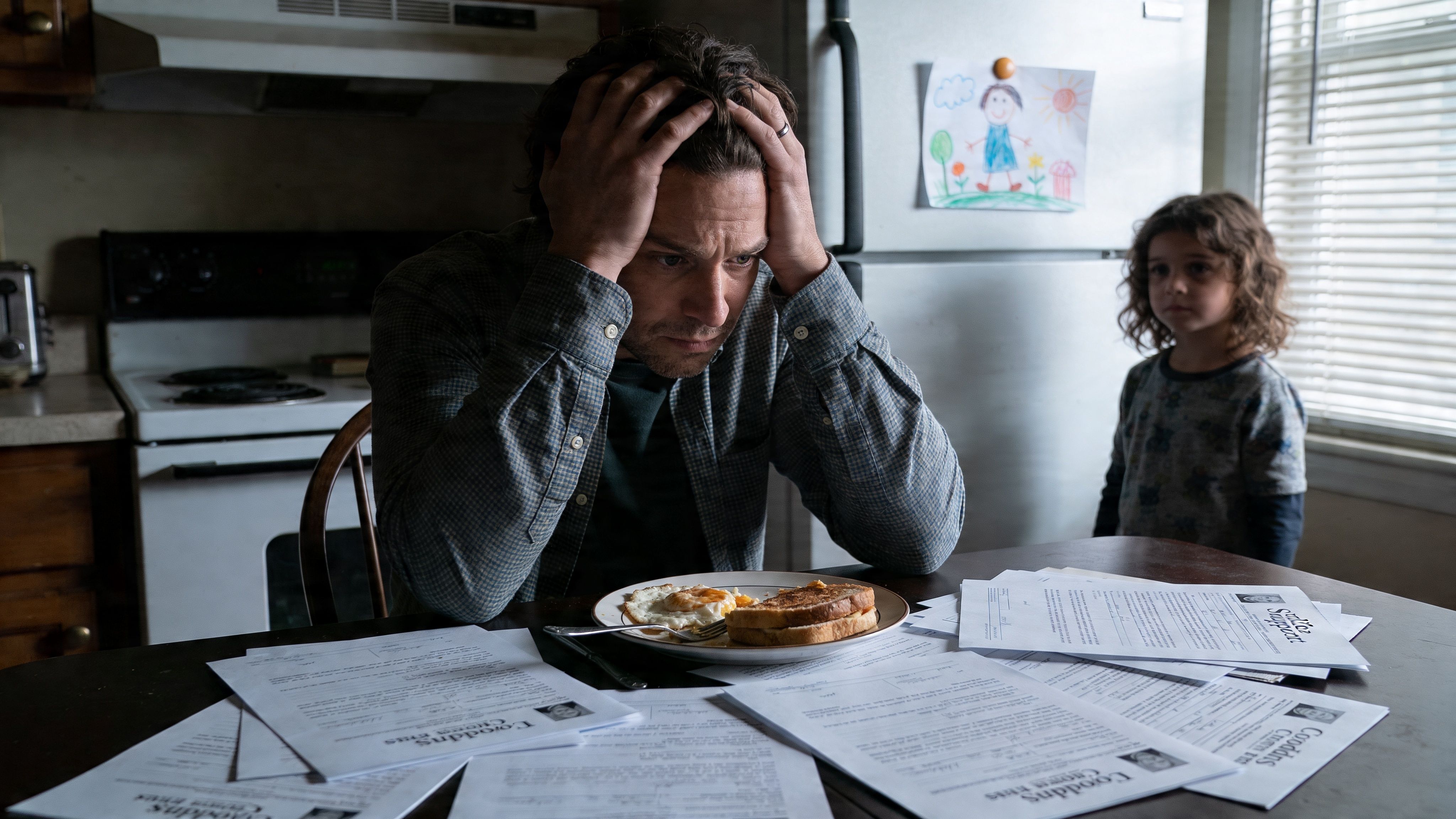A distressed father sits at a kitchen table surrounded by legal documents while his young child watches.