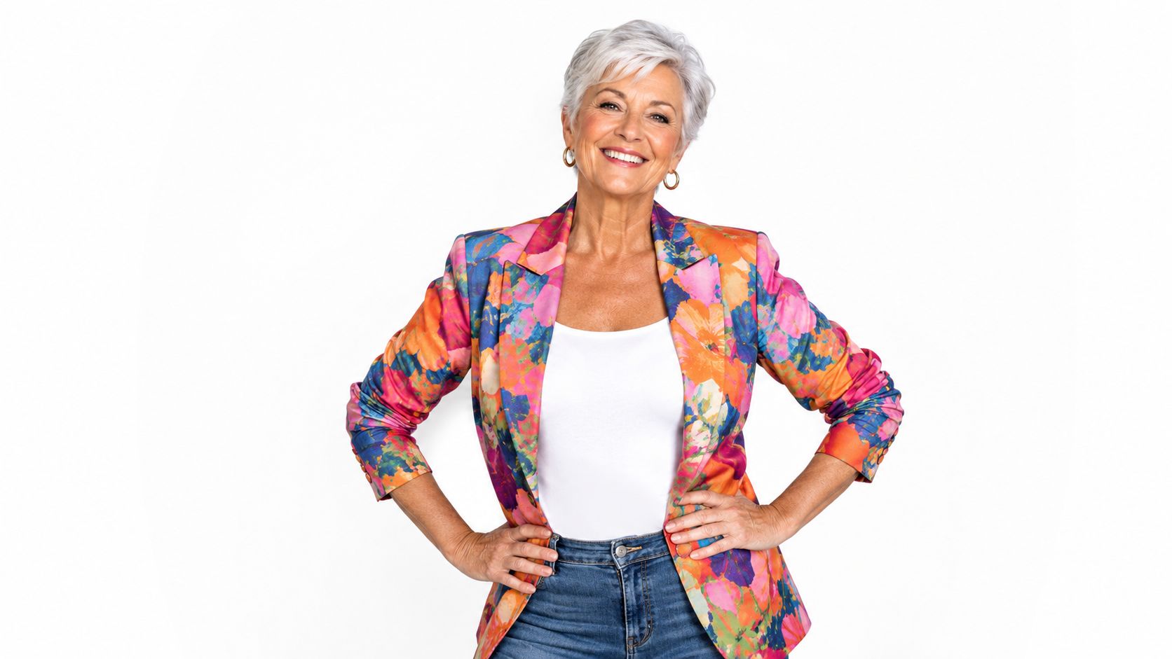 A happy senior woman with short grey hair wearing a colorful floral blazer and denim jeans.