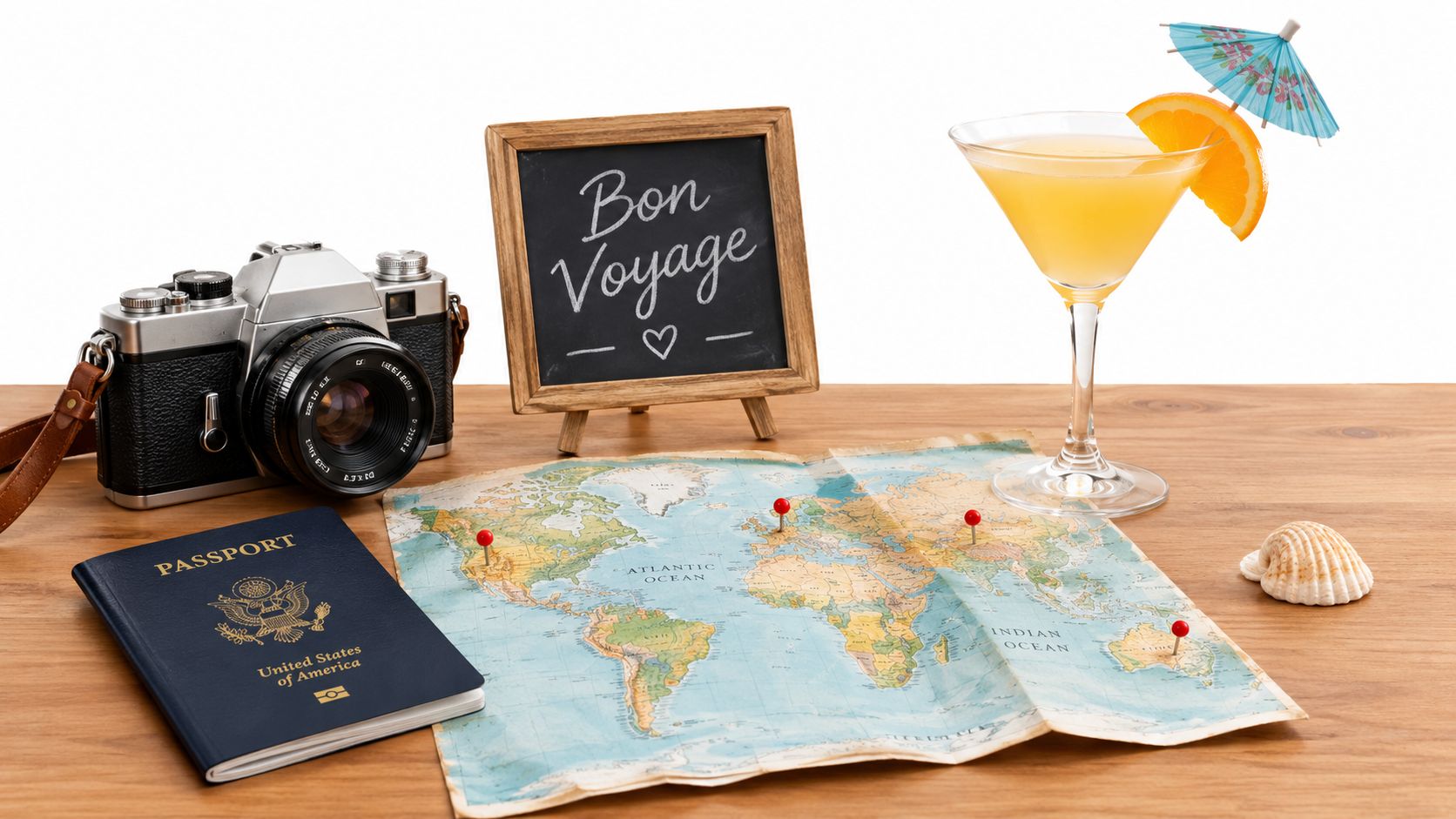A travel-themed desk setup featuring a camera, passport, world map, cocktail, and a Bon Voyage sign.