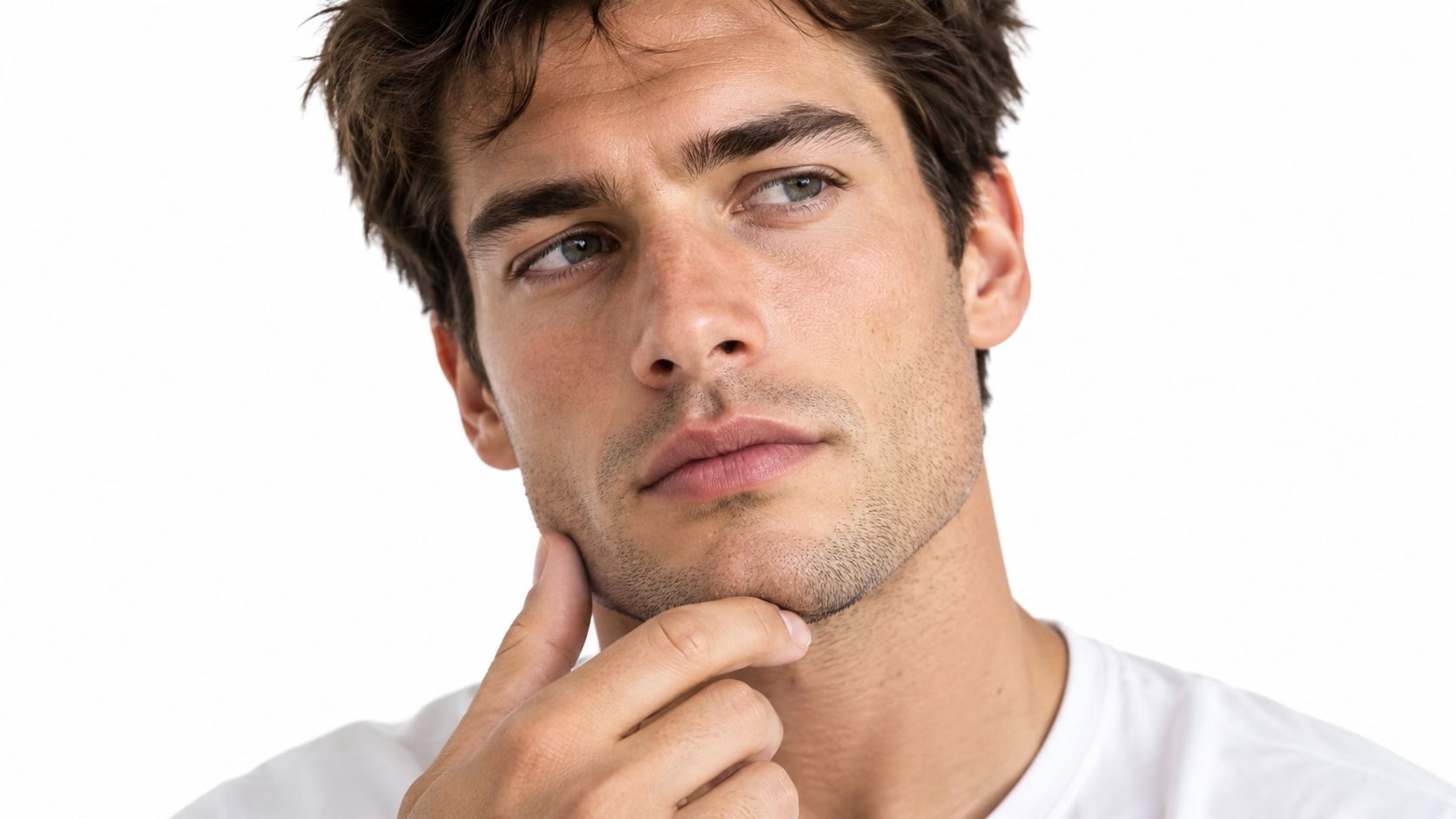A young man with clear skin touching his jawline while looking away in deep thought.