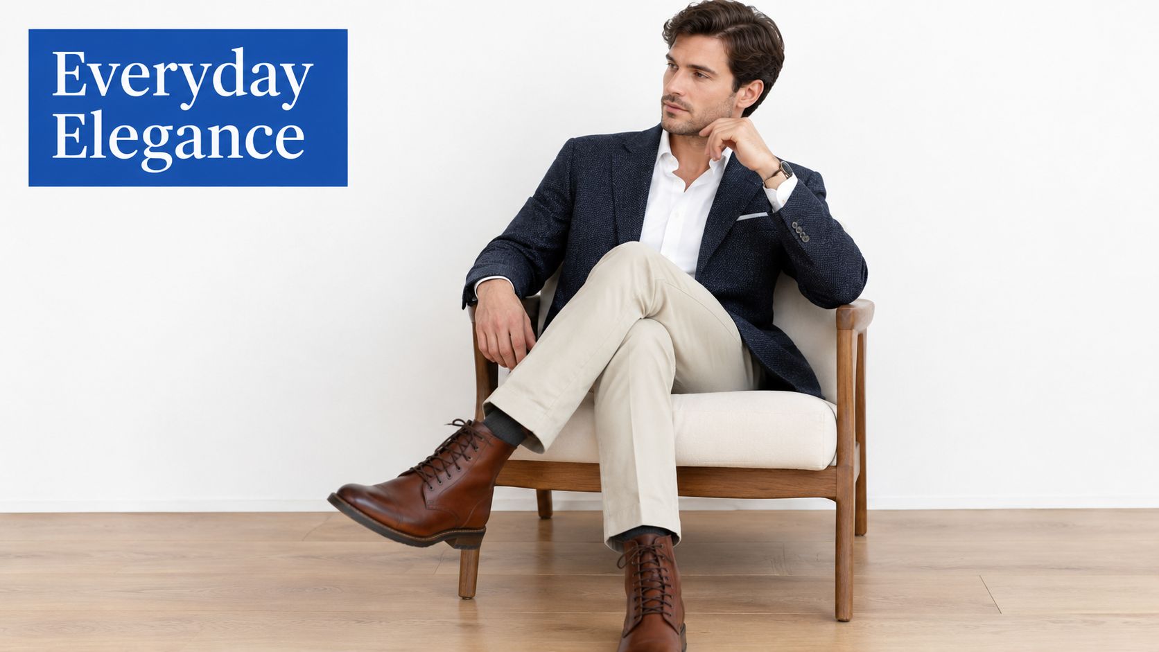 A stylish man in a blazer and khakis sits in a chair wearing brown leather boots.