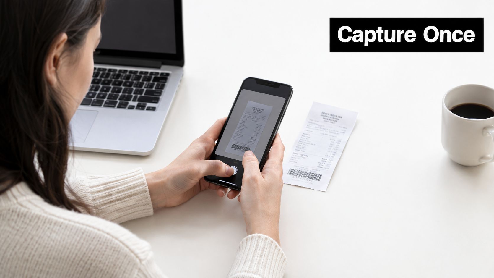 A person using a smartphone to scan a paper receipt while sitting at a desk with a laptop.