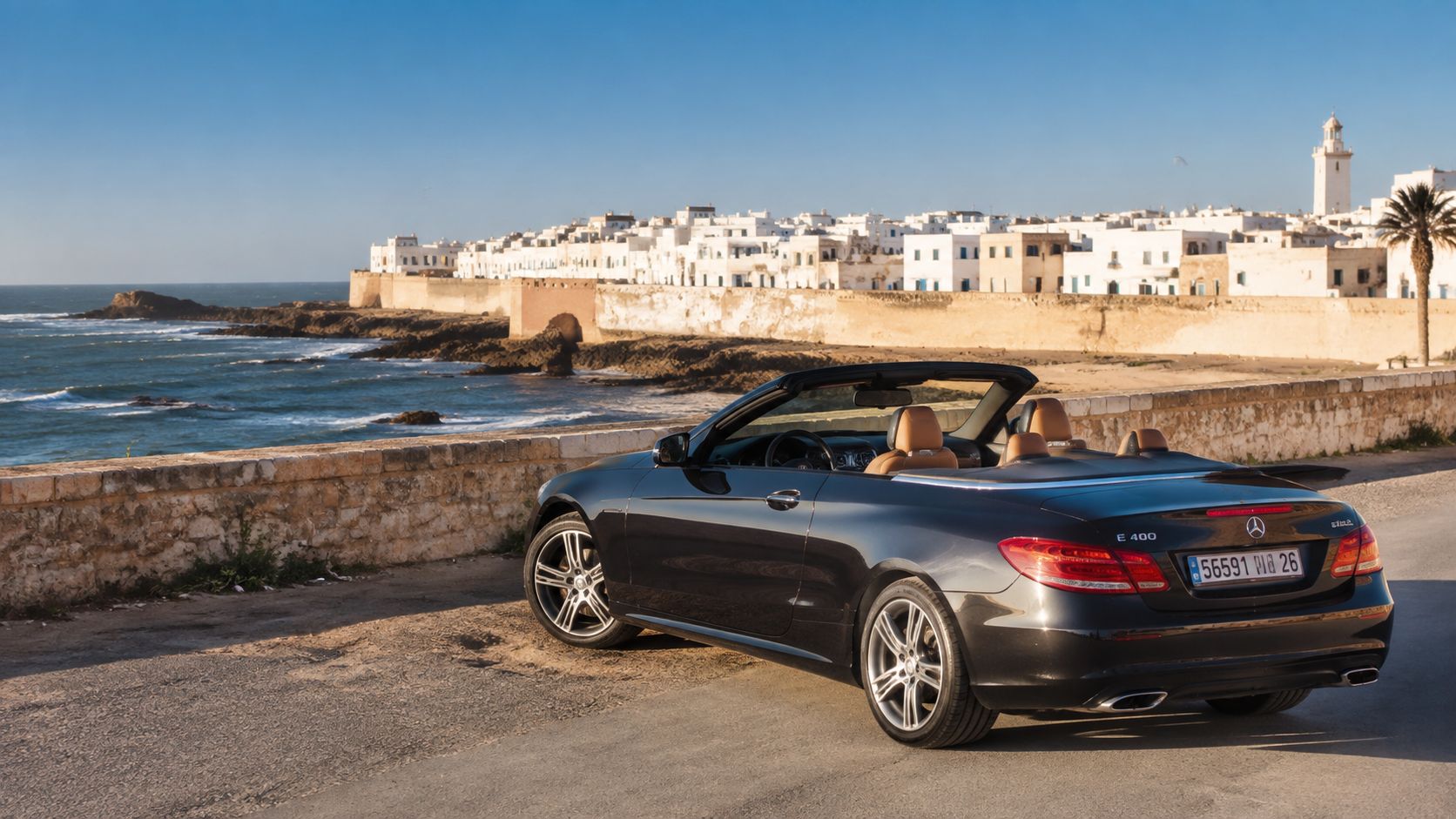 Une voiture décapotable noire stationnée sur le bord de mer avec une ville historique en arrière-plan.