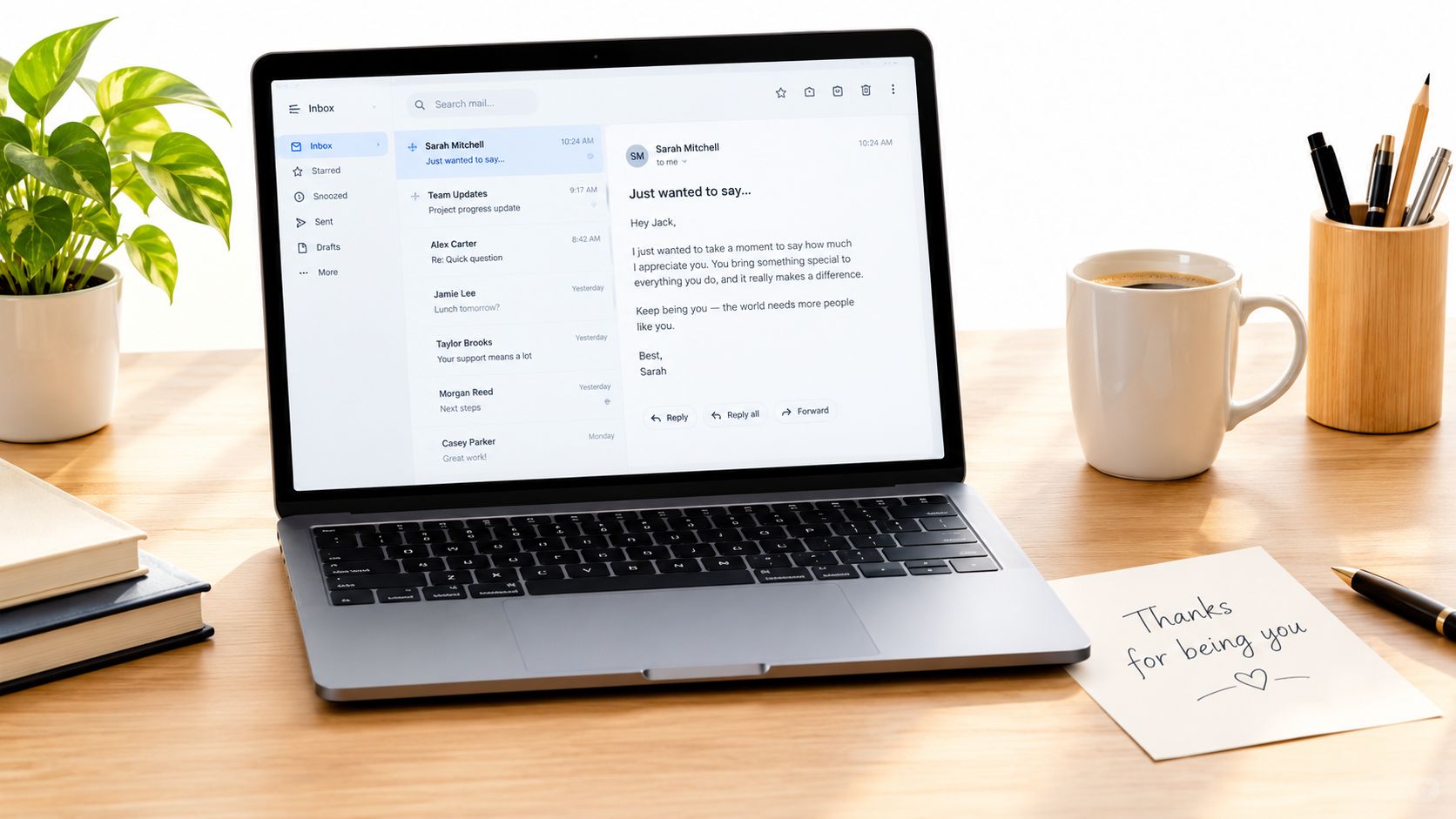 A modern laptop on a wooden desk displaying an email inbox, with a plant and coffee mug nearby.