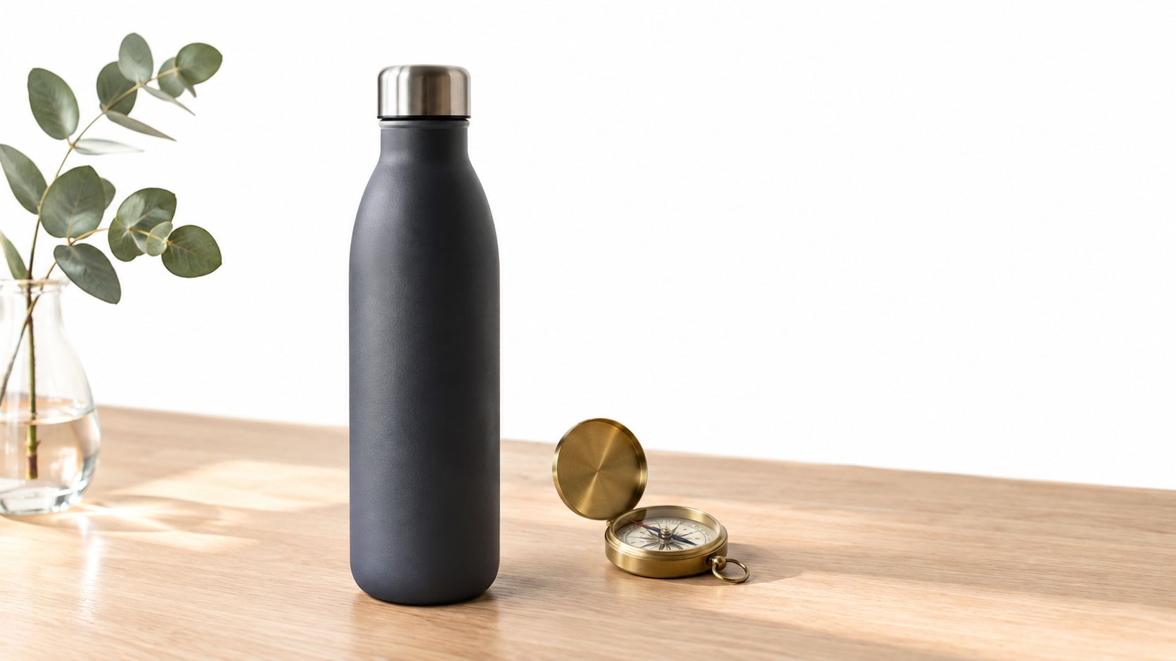 A matte dark grey water bottle standing next to a vintage brass compass on a wooden table.