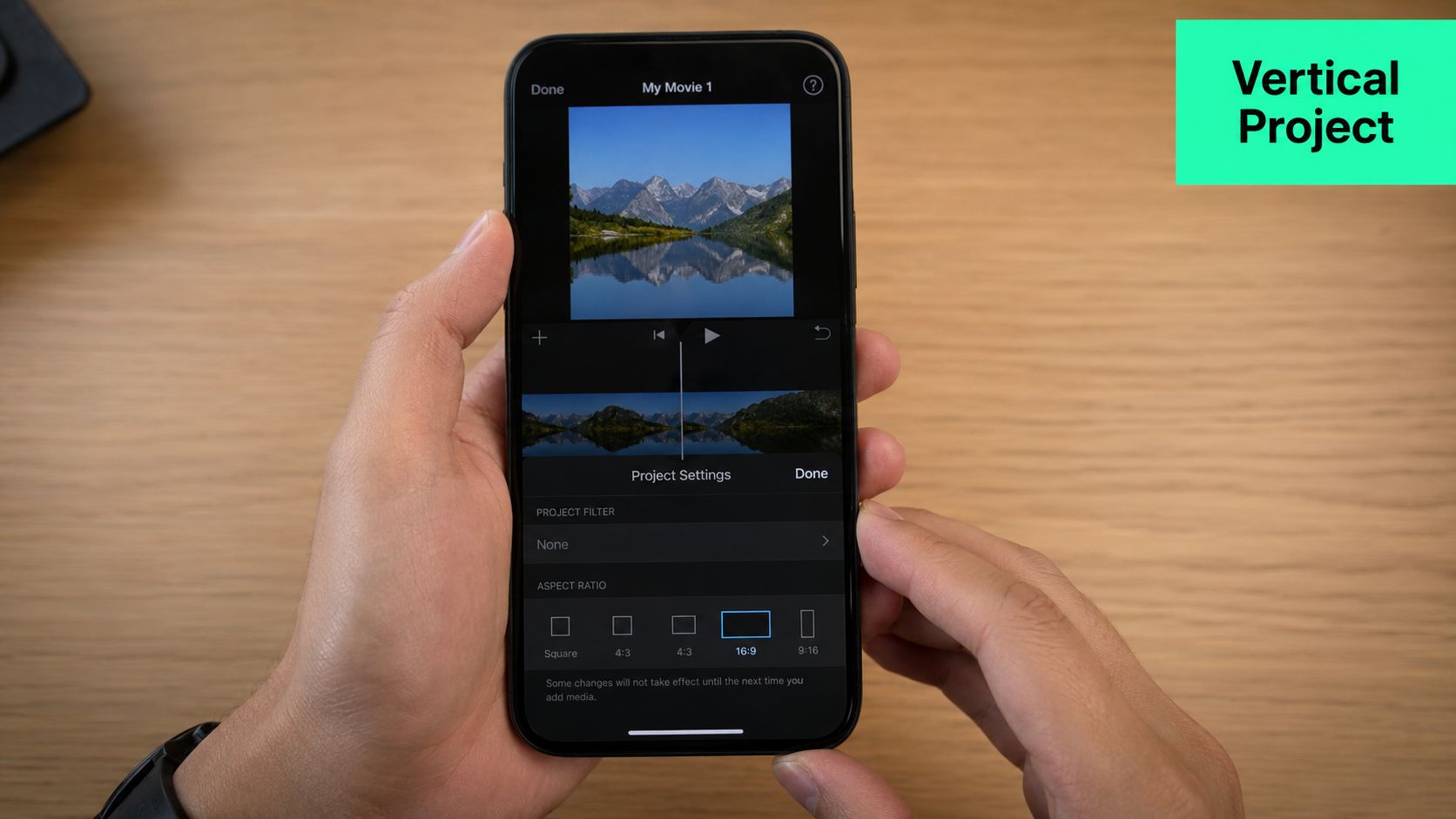 A close-up of a person holding an iPhone displaying the iMovie project settings and aspect ratio menu.