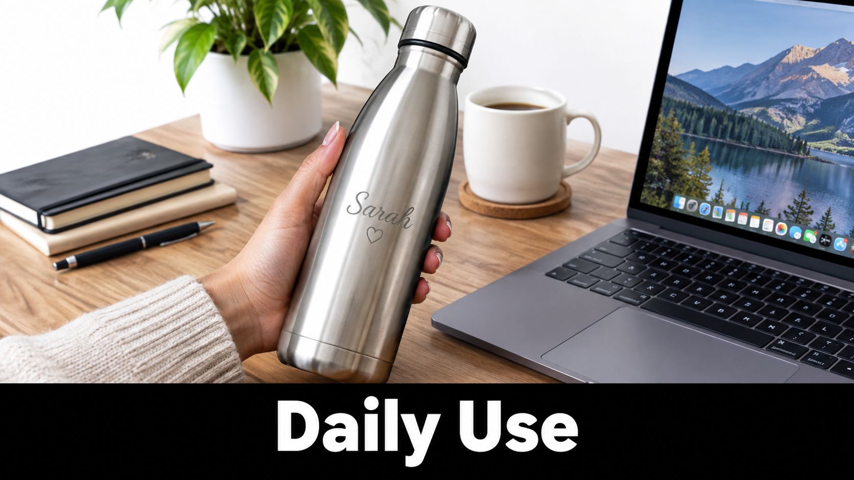 A person holding a personalized stainless steel water bottle on a wooden desk near a laptop.