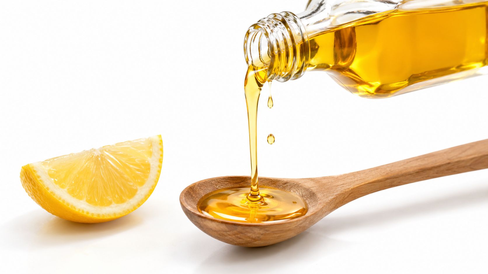 Golden oil being poured from a glass bottle into a wooden spoon next to a lemon wedge.